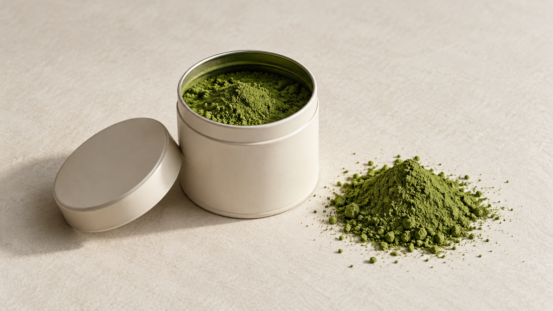 An open ivory tin of matcha beside a small mound of green tea powder on a textured cream background.
