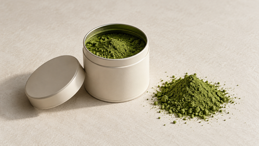 An open ivory tin of matcha beside a small mound of green tea powder on a textured cream background.