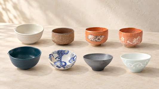 Eight ceramic matcha bowls in different shapes, colors, and finishes arranged on an ivory tabletop.